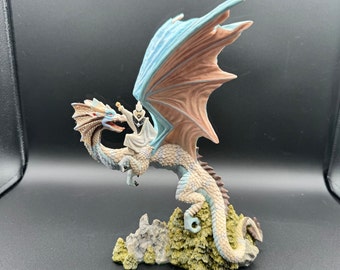 May include: A white figurine of a wizard riding a blue and brown dragon with a rocky base.