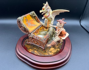 May include: A white dragon with a yellow belly and a small, brown, winged creature with pointed ears are standing on a wooden base. The dragon is looking at a small, ornate, wooden chest with a golden interior. The chest is open and appears to be empty.