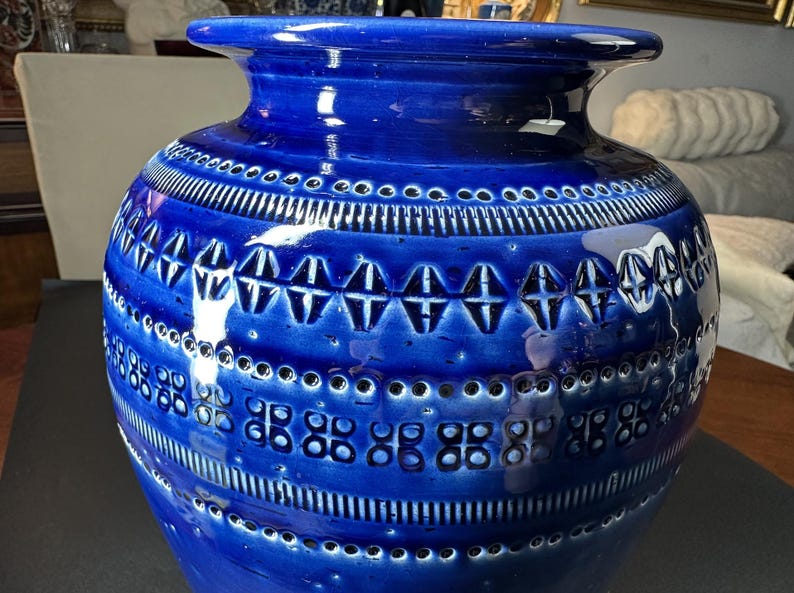 May include: A large, cobalt blue ceramic vase with a glossy finish. The vase features intricate geometric patterns, including rows of small circles, vertical lines, and diamond-shaped designs. The vase has a wide base and a flared rim, making it a decorative home accent.