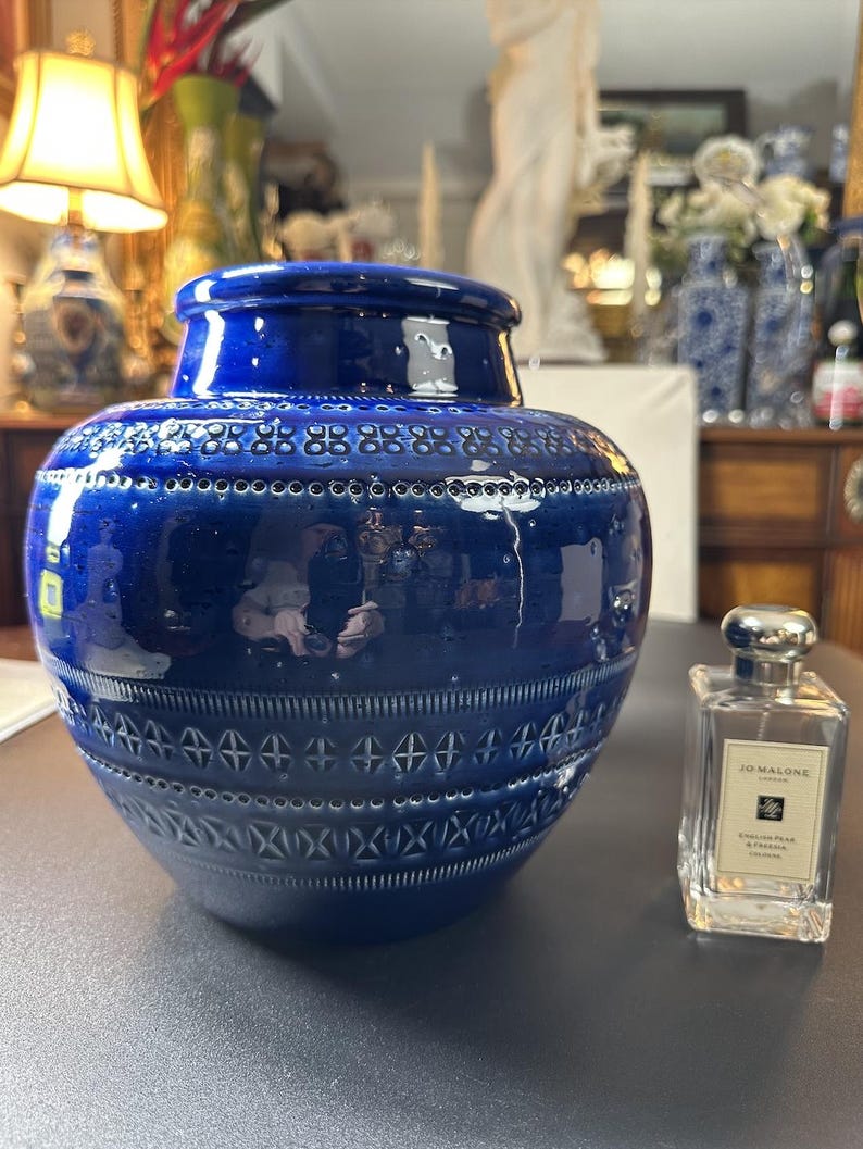 May include: A large, glossy, cobalt blue ceramic vase with a decorative pattern of small circles and geometric shapes. A Jo Malone cologne bottle is next to the vase.