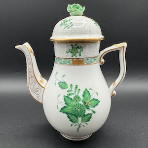 Puede incluir: Una cafetera de porcelana blanca con un diseño floral verde y dorado. La cafetera tiene una tapa con un diseño floral verde y un borde dorado. La cafetera está sobre un fondo negro.