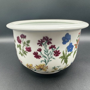 May include: White ceramic planter with a floral design featuring red, pink, blue, and yellow flowers. The planter has a wide rim and a rounded base.
