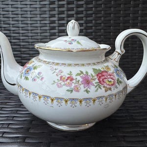 May include: A white porcelain teapot featuring a colorful floral pattern with pink, red, and blue flowers. The teapot has a matching lid with a small handle, a curved spout, and a looped handle, all accented with delicate gold trim along the edges.