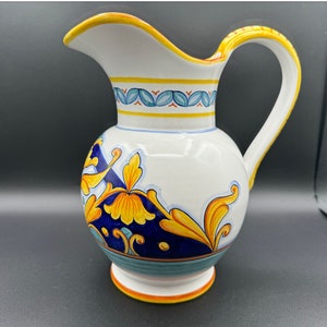May include: A white ceramic pitcher with a blue and yellow floral design. The pitcher has a wide mouth and a handle.