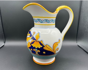 Può includere: Un'anfora in ceramica bianca con un motivo floreale blu e giallo. L'anfora ha un'ampia apertura e un manico.