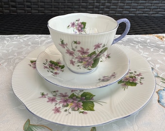 May include: A white teacup and saucer set with a delicate floral pattern of purple violets and green leaves. The teacup has a light blue handle and the saucer has a scalloped edge.