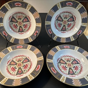 May include: A set of four round ceramic soup bowls featuring a white centre with a floral medallion pattern in pink, green, and dark blue. The wide rims are decorated with a dark blue grid pattern, gold accents, and repeating floral motifs.