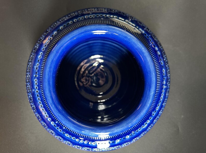 May include: A top-down view of a deep blue ceramic bowl. The bowl has a wide, textured rim with a repeating pattern of small, white dots. The interior is a darker shade of blue, with a glossy finish. The bowl's design is simple and elegant.