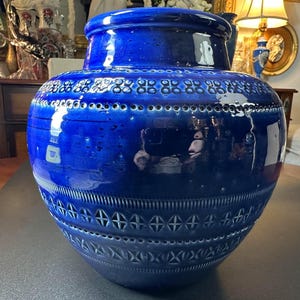 May include: A large, cobalt blue ceramic vase with a glossy finish. The vase features intricate geometric patterns around the neck and body, including rows of small circles and cross-like shapes. The vase has a wide, rounded body and a slightly flared rim.