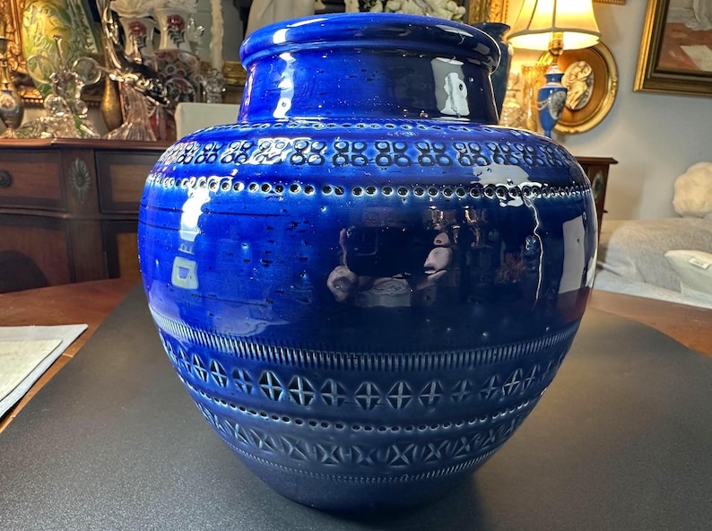 May include: A large, cobalt blue ceramic vase with a glossy finish. The vase features intricate geometric patterns around the neck and body, including rows of small circles and cross-like shapes. The vase has a wide, rounded body and a slightly flared rim.