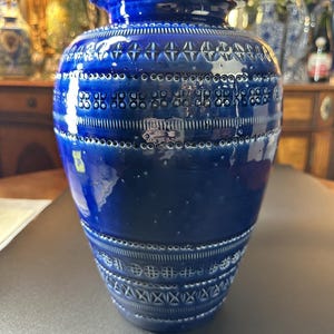 May include: A cobalt blue ceramic vase with a glossy finish. The vase features a series of textured, geometric patterns around the body, including rows of circles and cross-like designs. The vase has a wide base and a narrower neck.
