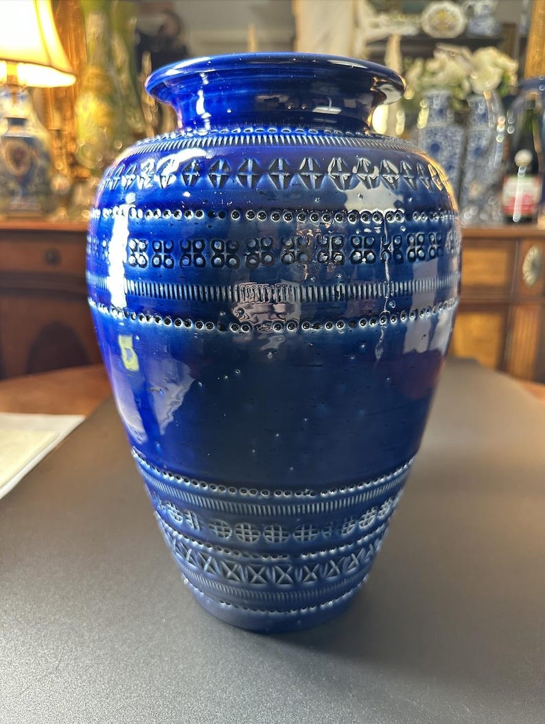 May include: A cobalt blue ceramic vase with a glossy finish. The vase features a series of textured, geometric patterns around the body, including rows of circles and cross-like designs. The vase has a wide base and a narrower neck.
