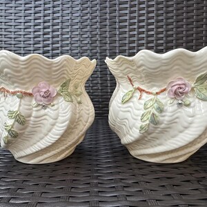 Könnte beinhalten: Zwei cremefarbene Keramik-Blumentöpfe mit gewelltem Design und floralen Akzenten. Jeder Topf hat eine rosa Rose, grüne Blätter und einen braunen Stiel. Geeignet für den Innen- oder Außenbereich.