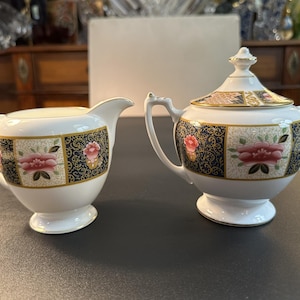 Könnte beinhalten: Ein weißes Milchkännchen und eine Zuckerdose mit Deckel und zwei Henkeln aus Porzellan. Beide Teile zeigen ein dekoratives Muster aus abwechselnden Feldern mit dunkelblauen Blumenmotiven und rosa Blüten auf cremefarbenem Grund, verziert mit Goldrand.
