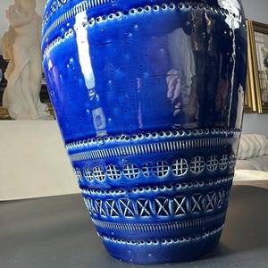 May include: A large, glossy blue ceramic vase with intricate patterns. The vase features a series of horizontal bands with geometric cutouts and textured designs. The vase is a decorative home accent, adding a pop of color and visual interest.