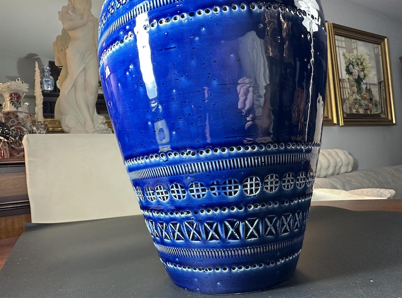 May include: A large, glossy blue ceramic vase with intricate patterns. The vase features a series of horizontal bands with geometric cutouts and textured designs. The vase is a decorative home accent, adding a pop of color and visual interest.