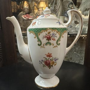 May include: White porcelain coffee pot with a floral design. The pot features a green and gold border, a curved handle, and a lid with a small finial. The floral pattern includes red, pink, and purple flowers. The base has a gold trim.