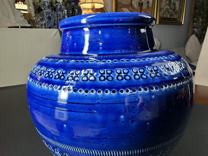 May include: A large, cobalt blue ceramic vase with a wide, rounded body and a slightly narrower neck. The vase features a series of decorative patterns, including rows of small circles and stylized geometric shapes. The glossy finish enhances the rich color.