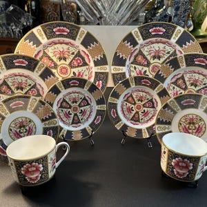 May include: A collection of fine china featuring two teacups and six plates of varying sizes. Each piece displays a dark blue and white background with intricate gold geometric patterns and vibrant pink floral motifs centered on the surface.