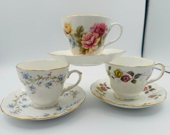 May include: Three vintage teacups and saucers with floral patterns. The cups are white with gold trim and feature different floral designs. The saucers are also white with gold trim and have matching floral patterns.