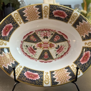 May include: An oval porcelain serving dish featuring a white center with a central floral medallion in pink, gold, and black. The wide rim displays alternating panels of dark blue with gold filigree, pink floral motifs, and gold scale patterns.