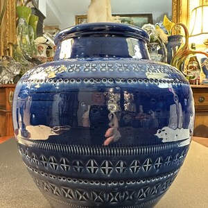 May include: A large, glossy, cobalt blue ceramic vase with a wide, rounded body and a slightly narrower neck. The vase features intricate embossed patterns, including geometric shapes and a series of small, evenly spaced holes near the top. The vase is a decorative home accent.