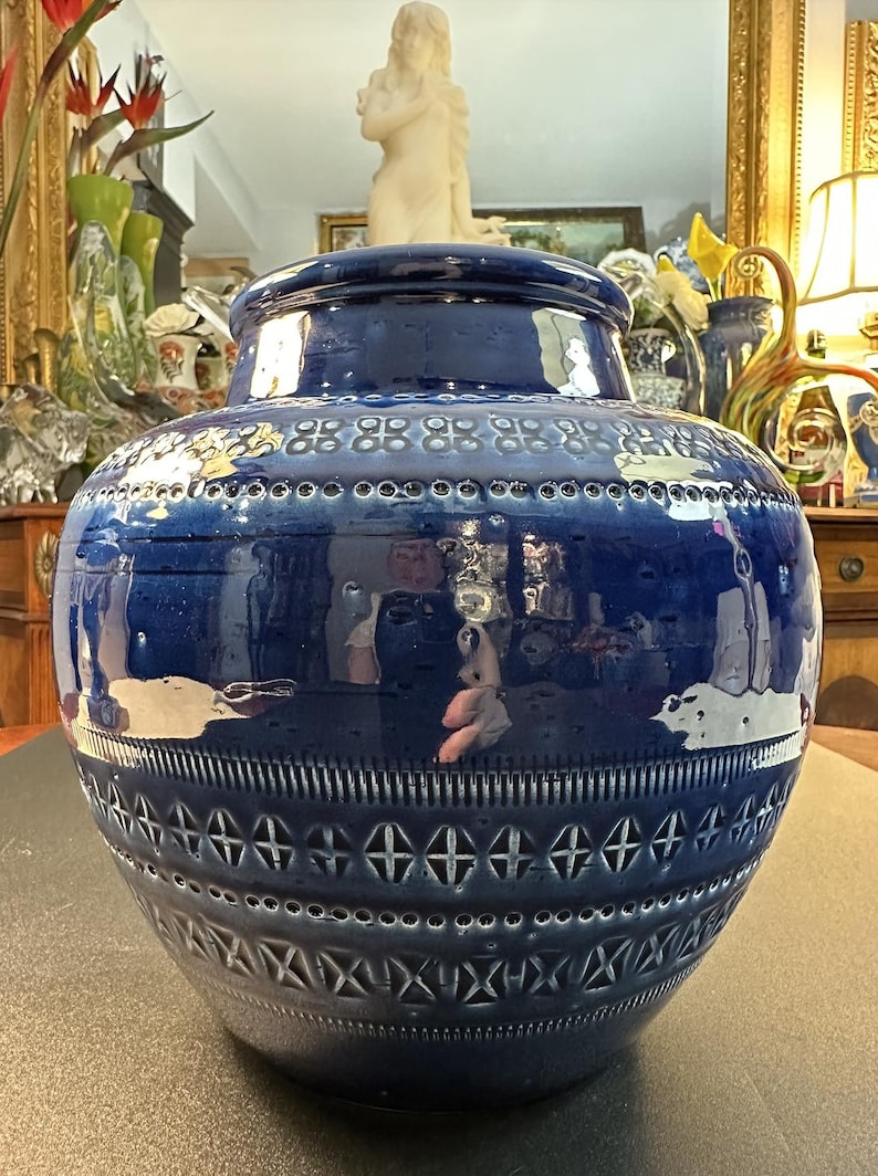 May include: A large, glossy, cobalt blue ceramic vase with a wide, rounded body and a slightly narrower neck. The vase features intricate embossed patterns, including geometric shapes and a series of small, evenly spaced holes near the top. The vase is a decorative home accent.
