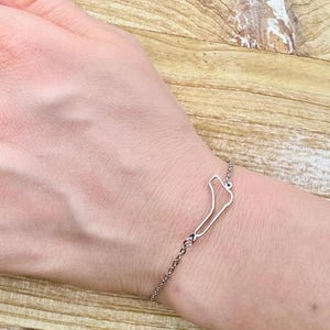 May include: A silver-colored bracelet featuring a minimalist outline design. The bracelet is a delicate chain with a pendant shaped like a stylized outline. The bracelet is worn on a wrist.