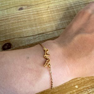Peut inclure: Un bracelet doré avec un motif abstrait minimaliste. Le bracelet est attaché à une chaîne fine et est porté au poignet. L'arrière-plan est une surface en bois.