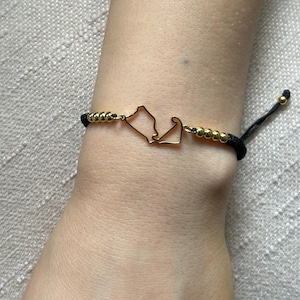 May include: Gold-coloured bracelet featuring a Cape Cod outline charm. The bracelet has a black cord with gold-coloured beads and an adjustable sliding knot closure. The charm is a detailed representation of the Cape Cod peninsula.