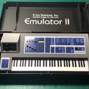 May include: A gray E-mu Systems, Inc. Emulator II synthesizer keyboard housed in a black carrying case. The control panel features blue sections with white text, a small yellow digital display, and various sliders and buttons. The keyboard has black and white keys.