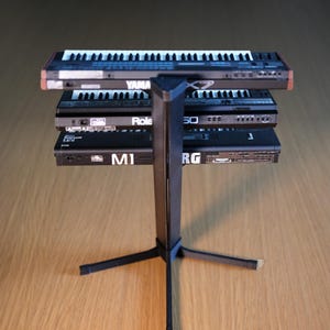 Op de afbeelding: Een zwarte metalen keyboardstandaard ondersteunt drie keyboards. Het bovenste keyboard is gemarkeerd met "YAMAHA", de middelste met "Role" en de onderste met "M1". De keyboards hebben witte en zwarte toetsen.
