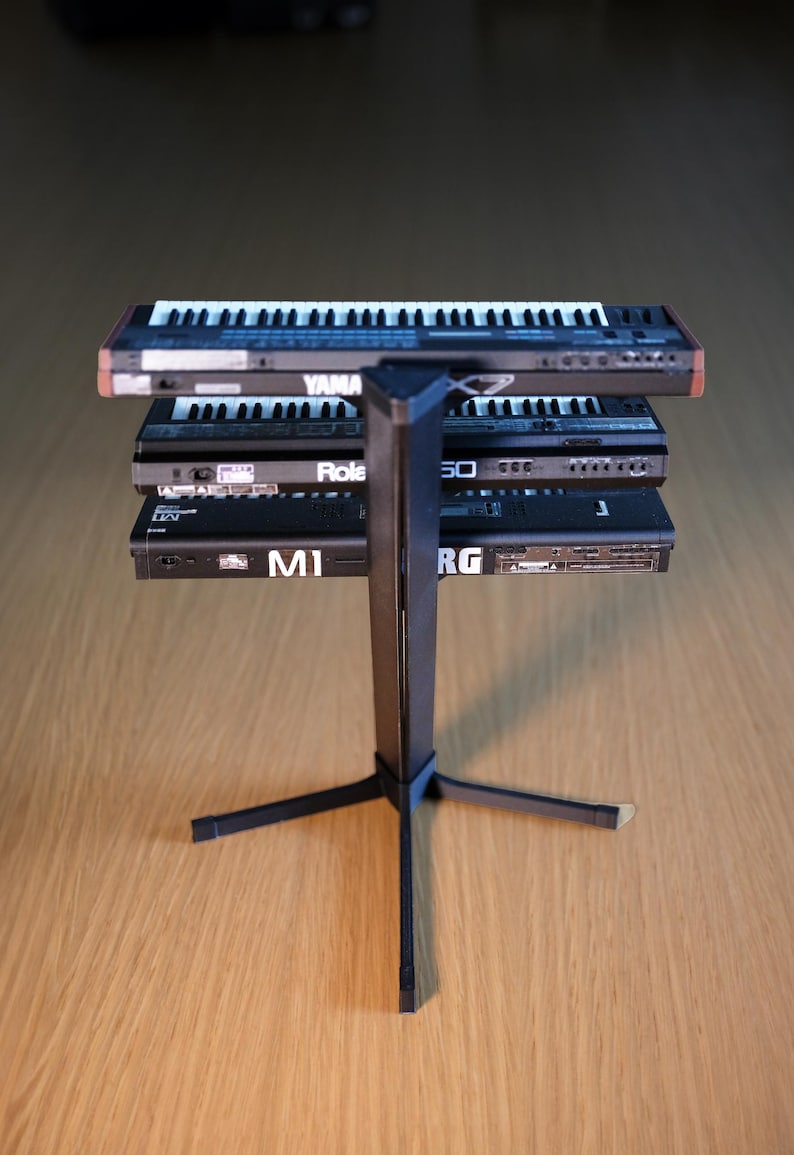 Op de afbeelding: Een zwarte metalen keyboardstandaard ondersteunt drie keyboards. Het bovenste keyboard is gemarkeerd met "YAMAHA", de middelste met "Role" en de onderste met "M1". De keyboards hebben witte en zwarte toetsen.