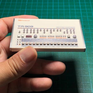 Puede incluir: Una réplica en miniatura blanca del Roland TR-909 Rhythm Composer que presenta un panel de control detallado con numerosos botones pequeños, perillas y etiquetas impresas. El dispositivo muestra el logotipo de Roland y el texto TR-909.