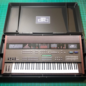 Może przedstawiać: Klawiatura syntezatora Yamaha DX1 w czarnej obudowie. Klawiatura ma ciemnoszarą obudowę z brązowym wykończeniem i pełnym zestawem białych i czarnych klawiszy. Obudowa jest otwarta, ukazując elementy sterujące i wyświetlacz instrumentu.