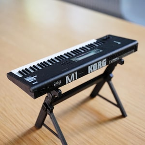 以下が含まれることがあります： 白と黒の鍵盤が付いた黒いM1 Korgシンセサイザー。黒いスタンドの上に置かれています。シンセサイザーには「M1 KORG」の文字が印刷されています。スタンドは含まれていません。