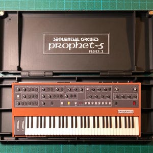 May include: A Sequential Circuits Prophet-5 Rev.1 synthesiser in a black case. The synthesiser has a burnt orange control panel with numerous knobs and switches, a keyboard, and the text "prophet-5" and "Sequential Circuits" on the panel.