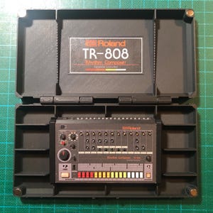 May include: A black Roland TR-808 Rhythm Composer, a vintage drum machine, is displayed inside a dark gray case. The case is open, revealing the machine's control panel with knobs, buttons, and a series of colored pads. The text "Roland TR-808 Rhythm Composer" is visible.