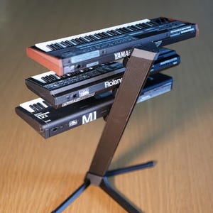 Op de afbeelding: Een zwarte keyboardstandaard met drie kleine synthesizers. De bovenste synth is gemarkeerd met Yamaha, de middelste met Roland en de onderste met MI. De keyboards hebben witte en zwarte toetsen.