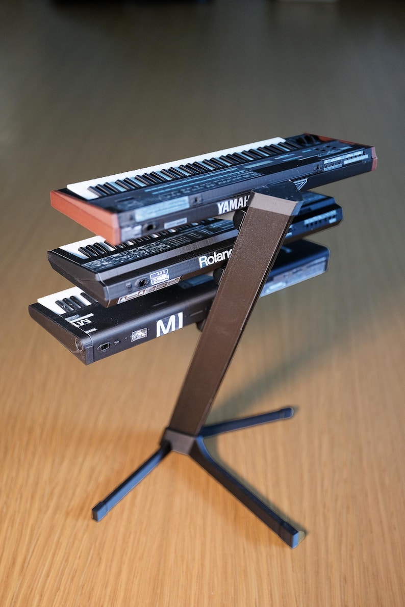 Op de afbeelding: Een zwarte keyboardstandaard met drie kleine synthesizers. De bovenste synth is gemarkeerd met Yamaha, de middelste met Roland en de onderste met MI. De keyboards hebben witte en zwarte toetsen.