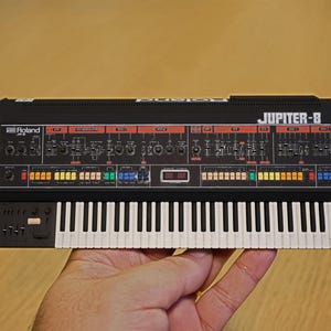 Puede incluir: Un sintetizador Roland Jupiter-8 negro y plateado con un teclado completo y numerosos botones y perillas de control. El texto "JUPITER-8" es visible en la parte superior derecha. El sintetizador se sostiene en una mano.