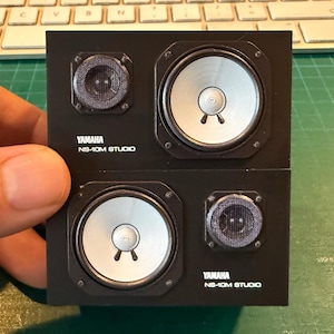 May include: A miniature black speaker unit featuring two white circular woofers and two black mesh tweeters. The front panel displays the white text Yamaha NS-10M STUDIO in two locations, reflecting the iconic design of professional studio monitors.