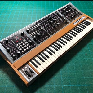 Puede incluir: Un modelo en miniatura de un sintetizador Memorymoog con chasis de color tostado, un panel de control negro con numerosos mandos y botones, y un teclado estándar en blanco y negro. El texto "memorymoog" es visible en la parte superior derecha.