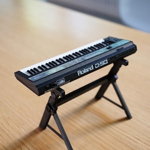 Op de afbeelding: Een zwart-wit miniatuur Roland D-50 synthesizer keyboard op een zwart statief. Het keyboard heeft witte en zwarte toetsen, met het Roland D-50 logo zichtbaar. Het statief is een driepoot ontwerp. De tekst "(STAND NOT INCLUDED)" staat onderaan.