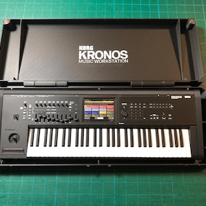 May include: A black Korg Kronos music workstation keyboard with a central color touchscreen, numerous control knobs, sliders, and a full-size piano-style keybed, housed within a matching black protective hard case with the brand name printed inside.
