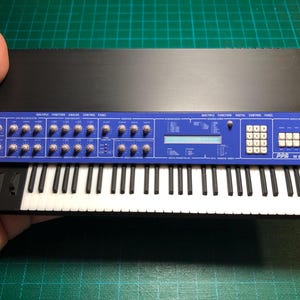 May include: A miniature black and blue synthesizer model featuring a keyboard with white and black keys, a control panel with numerous silver knobs, a digital display screen, a numeric keypad, and the text PPG wave 2.3 printed on the right side.