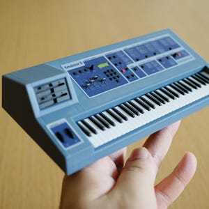 May include: A light blue synthesizer with black and white keys, labeled "Emulator II." The instrument has various buttons, knobs, and a display screen. The keyboard is held in a hand.