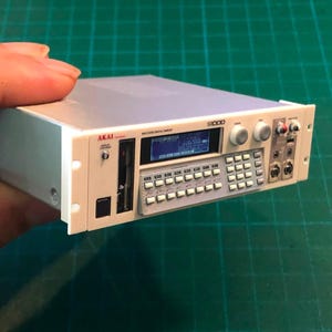Puede incluir: Una réplica en miniatura del sampler digital Akai S1000 con acabado en gris claro. El dispositivo cuenta con una pequeña pantalla digital azul, un teclado numérico, varios mandos de control y puertos de entrada en el panel frontal.