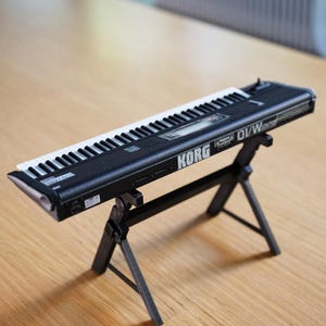 Op de afbeelding: Een zwart Korg DV/W synthesizer keyboard met witte en zwarte toetsen, op een zwarte standaard. Het keyboard staat op een lichtbruin houten oppervlak. De teksten "KORG" en "DV/W" zijn zichtbaar. De tekst "(STAND NOT INCLUDED)" staat onderaan.