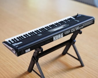 May include: A black Korg DV/W synth keyboard with white and black keys, resting on a black stand. The keyboard is on a light brown wooden surface. The text "KORG" and "DV/W" are visible on the keyboard. The text "(STAND NOT INCLUDED)" is at the bottom.